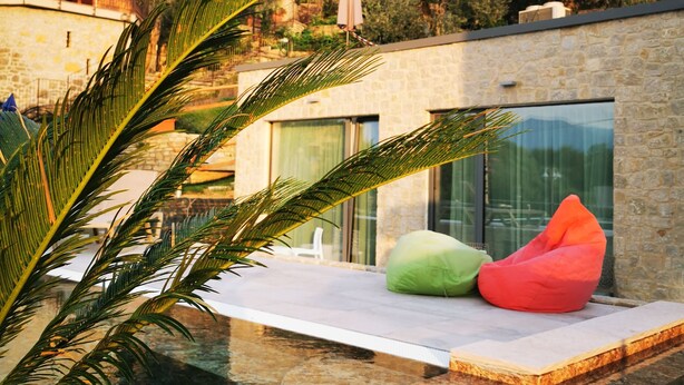A modern outdoor pool area with a stone deck, a glass-enclosed seating area with colorful bean bags, and a view of surrounding buildings and greenery. - Hotel San Marco (Torri del Benaco)