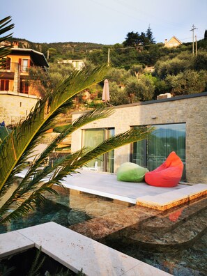 A modern outdoor pool area with a stone deck, a glass-enclosed seating area with colorful bean bags, and a view of surrounding buildings and greenery.