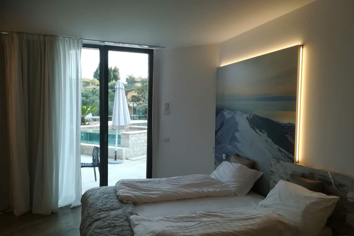 Ein Hotelzimmer mit Bett, einem Gemälde an der Wand und Blick auf einen Außenbereich mit Pool und Sonnenschirmen.