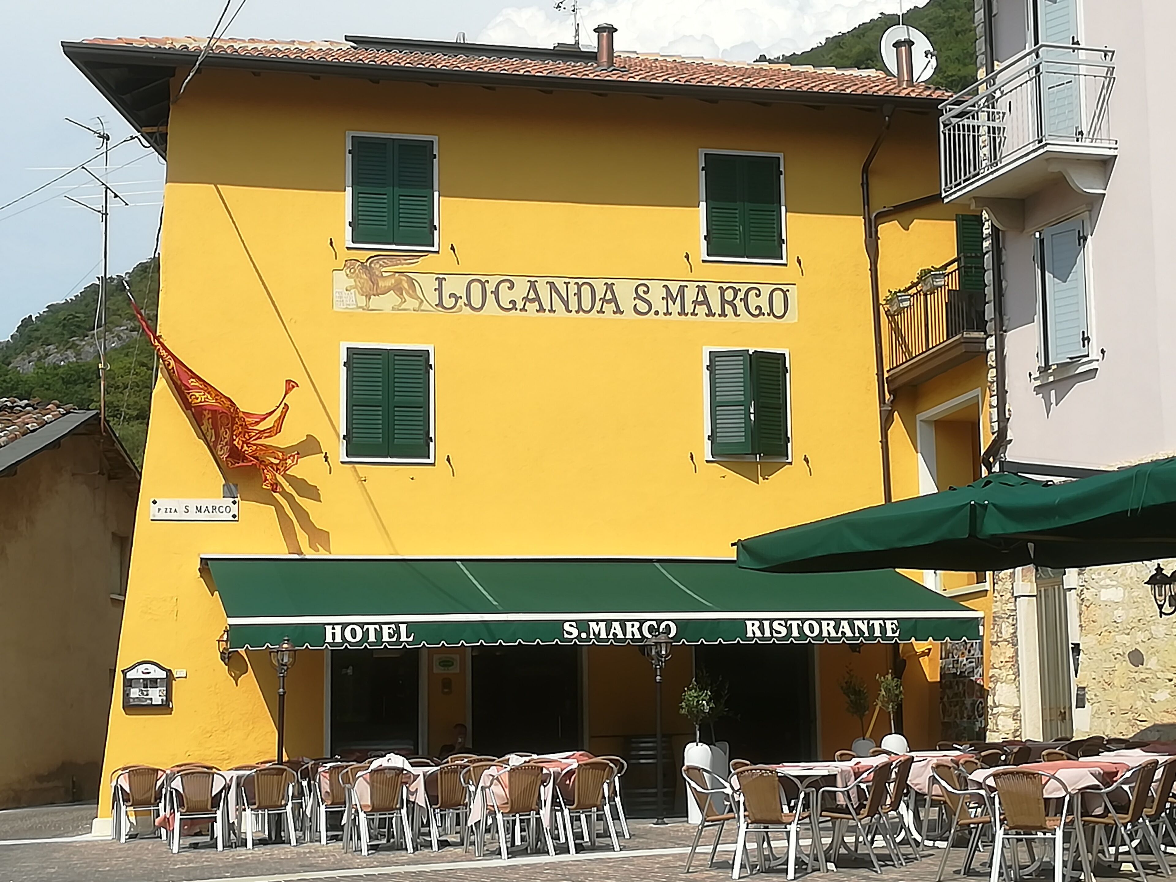 Ein gelbes Gebäude mit der Aufschrift „Hotel S. Marco“ und „Ristorante“, das ein Restaurant kennzeichnet. Vor dem Gebäude befinden sich Tische und Stühle im Freien.