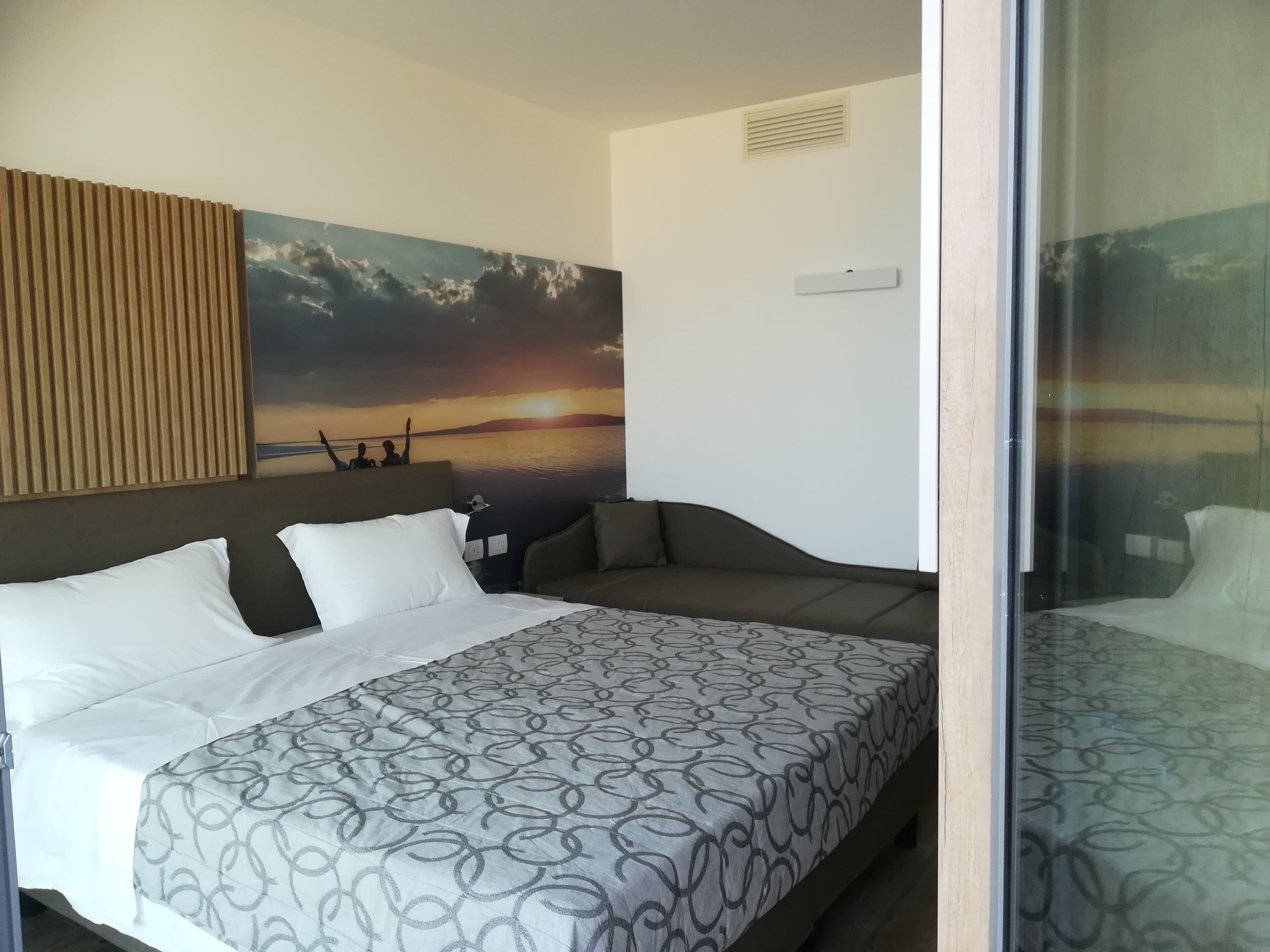 Habitación de hotel con cama, sofá, una obra de arte en la pared que representa un atardecer y ventana con vistas.