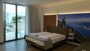 Quarto de hotel com uma cama, vista para uma piscina e montanhas, e uma pintura grande de um caminhante.