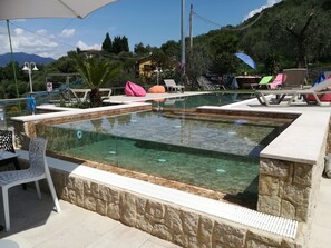 A clear pool with stone walls, surrounded by lounge chairs and umbrellas, with a mountainous backdrop.