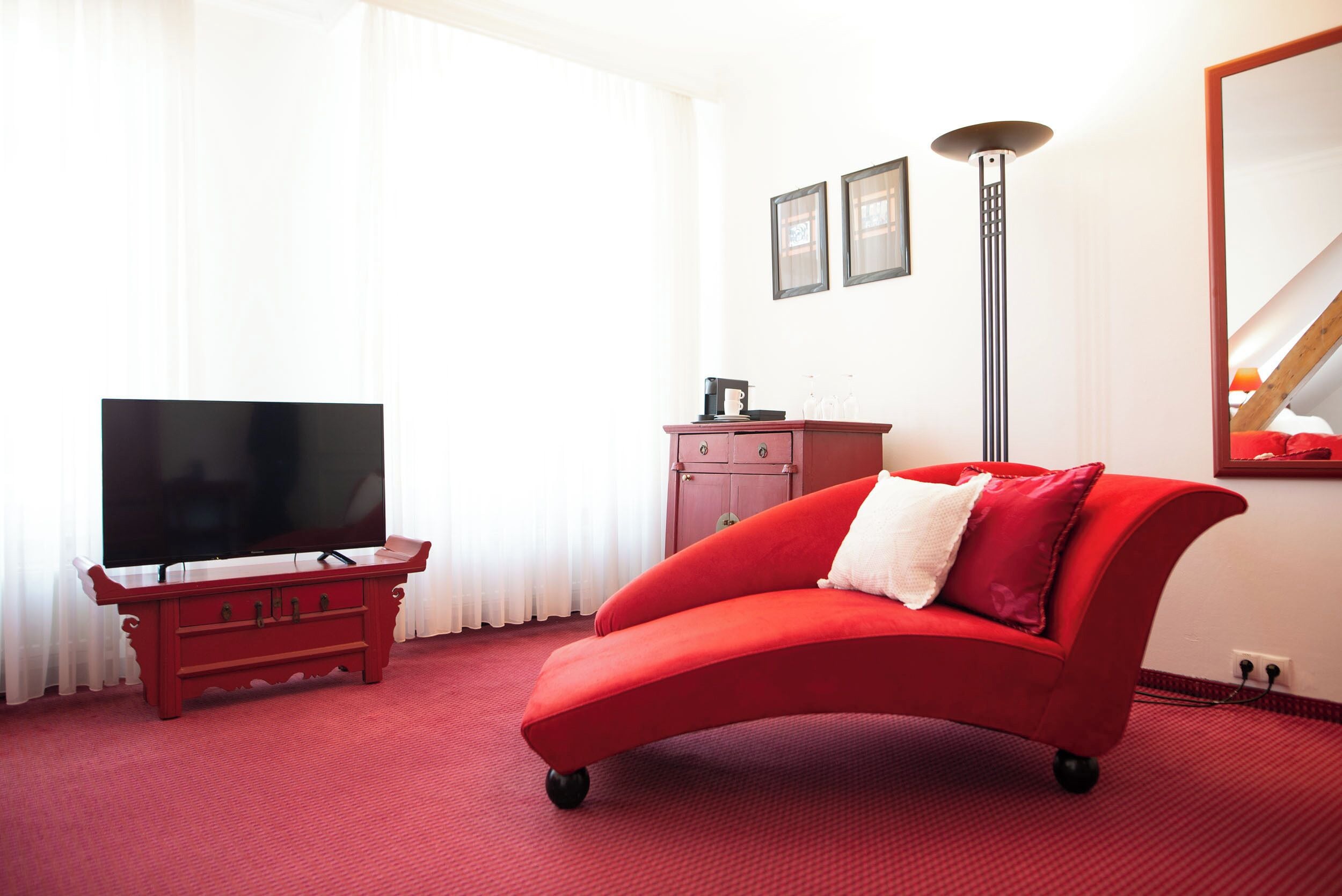 A red sofa, a television on a stand, a cabinet, and a tall floor lamp in a room with red carpet.