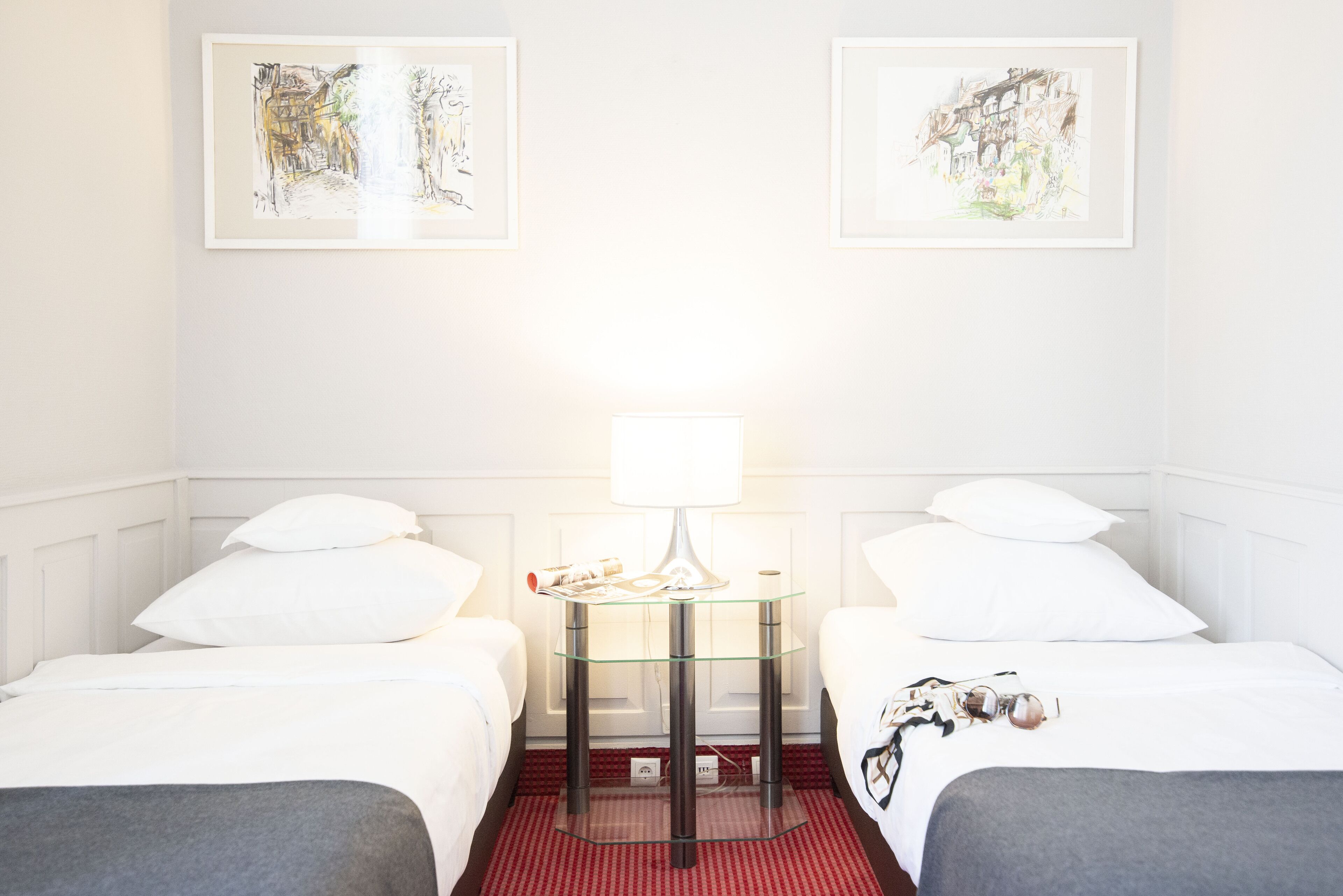 A hotel room with two beds, a glass table with a lamp, and two framed artworks on the wall.