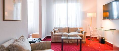 Habitación de hotel con alfombra roja, dos sofás, una mesita de café, un televisor y una lámpara.
