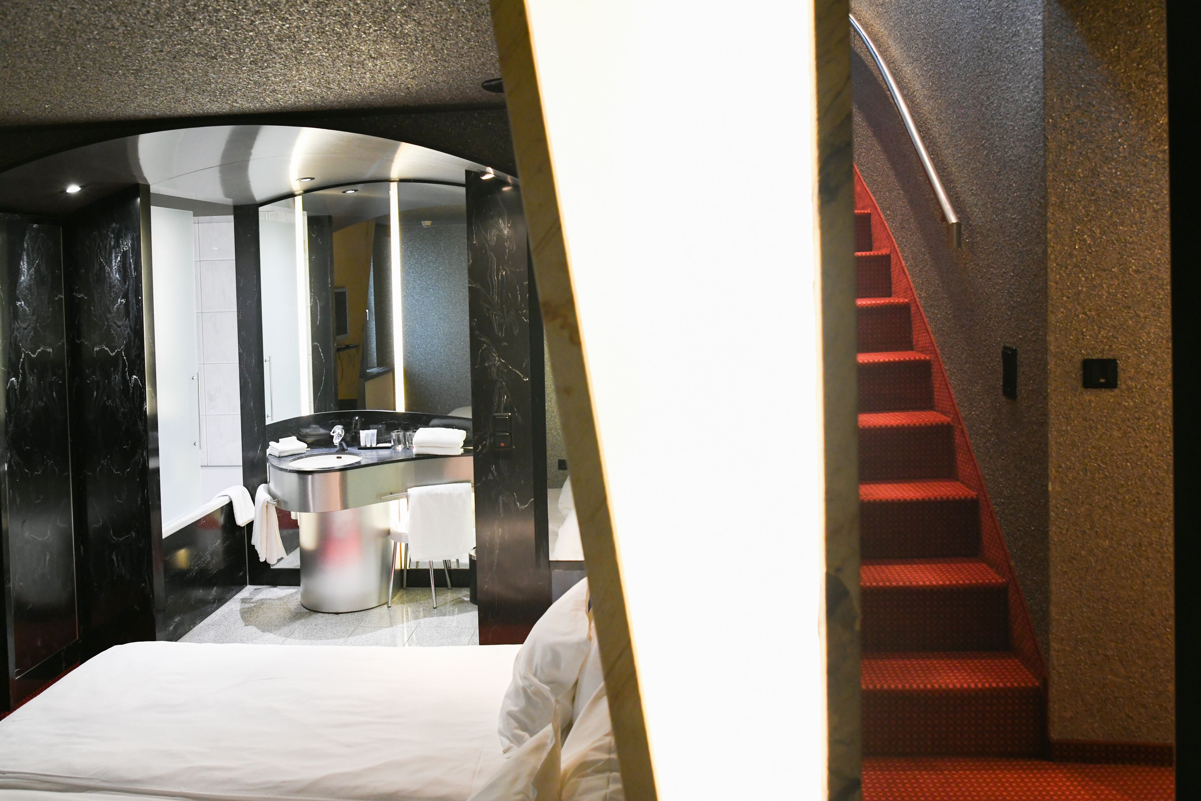 Une chambre d’hôtel avec un lit, une salle de bain dotée d’un lavabo et d’un miroir, et un escalier avec des marches rouges.