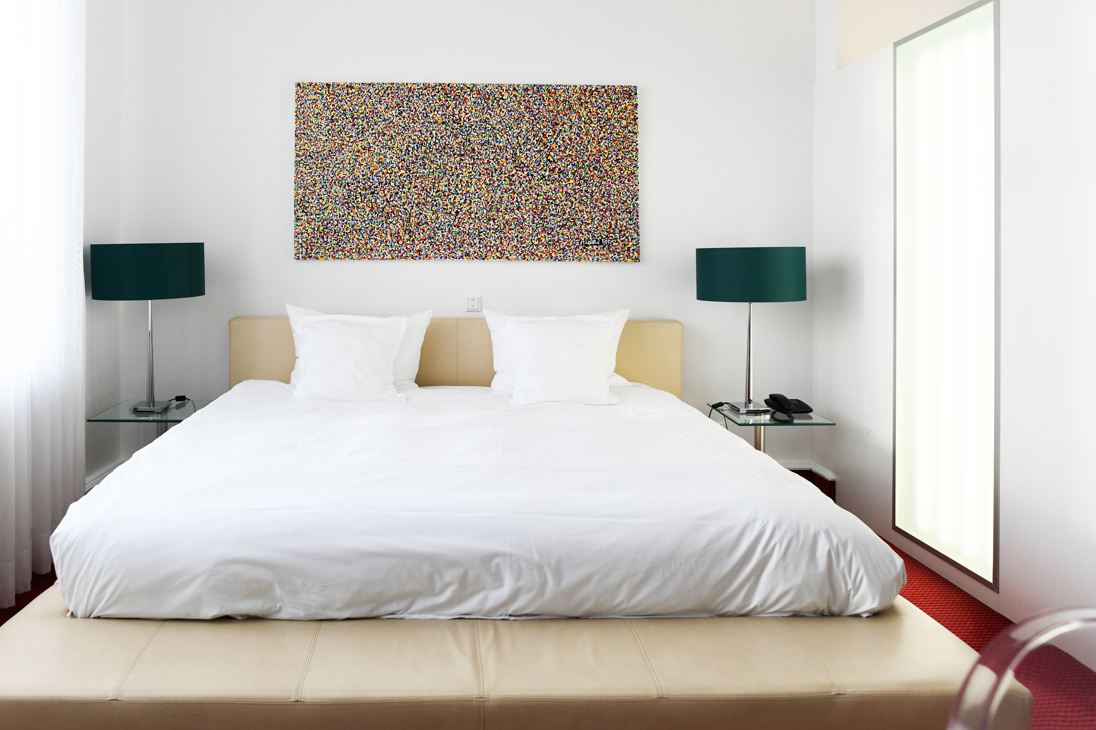Una habitación de hotel moderna con una cama grande, dos mesitas de noche con lámparas, un cuadro abstracto y colorido en la pared, y un ventanal que deja entrar luz natural.