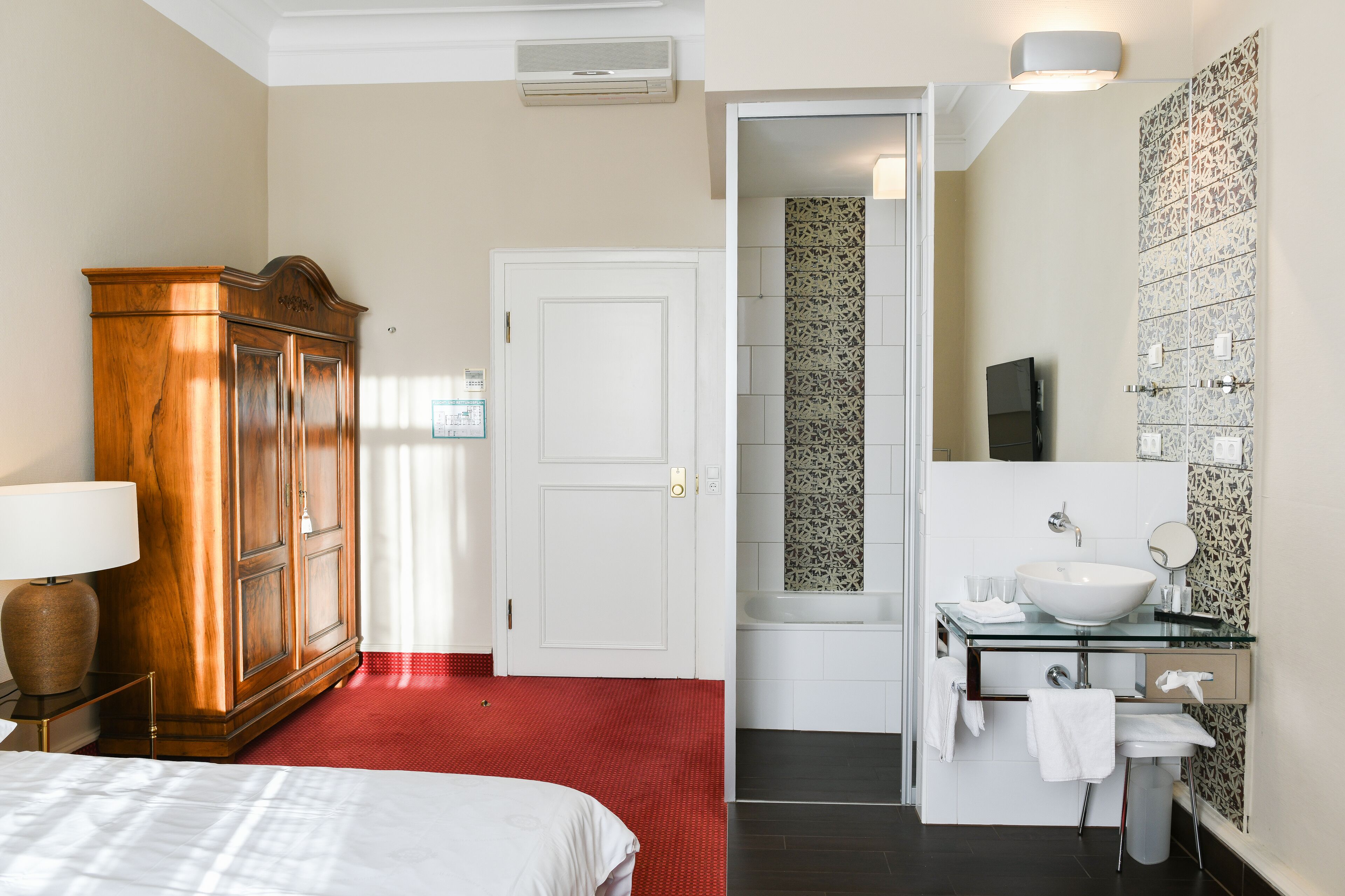 Ein Hotelzimmer mit Holzschrank, einem Bett mit weißer Bettwäsche, einem roten Teppich, einem Badezimmer mit Waschbecken aus Glas und einer Badewanne.