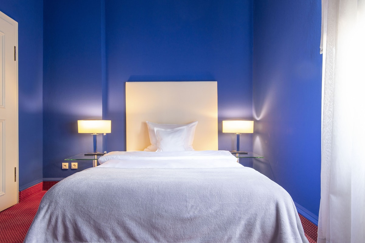 Quarto de hotel com uma cama grande, duas luminárias de cabeceira e uma parede azul.