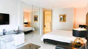 A modern hotel room with a bed, bedside table, lamp, and a glass desk with a chair.