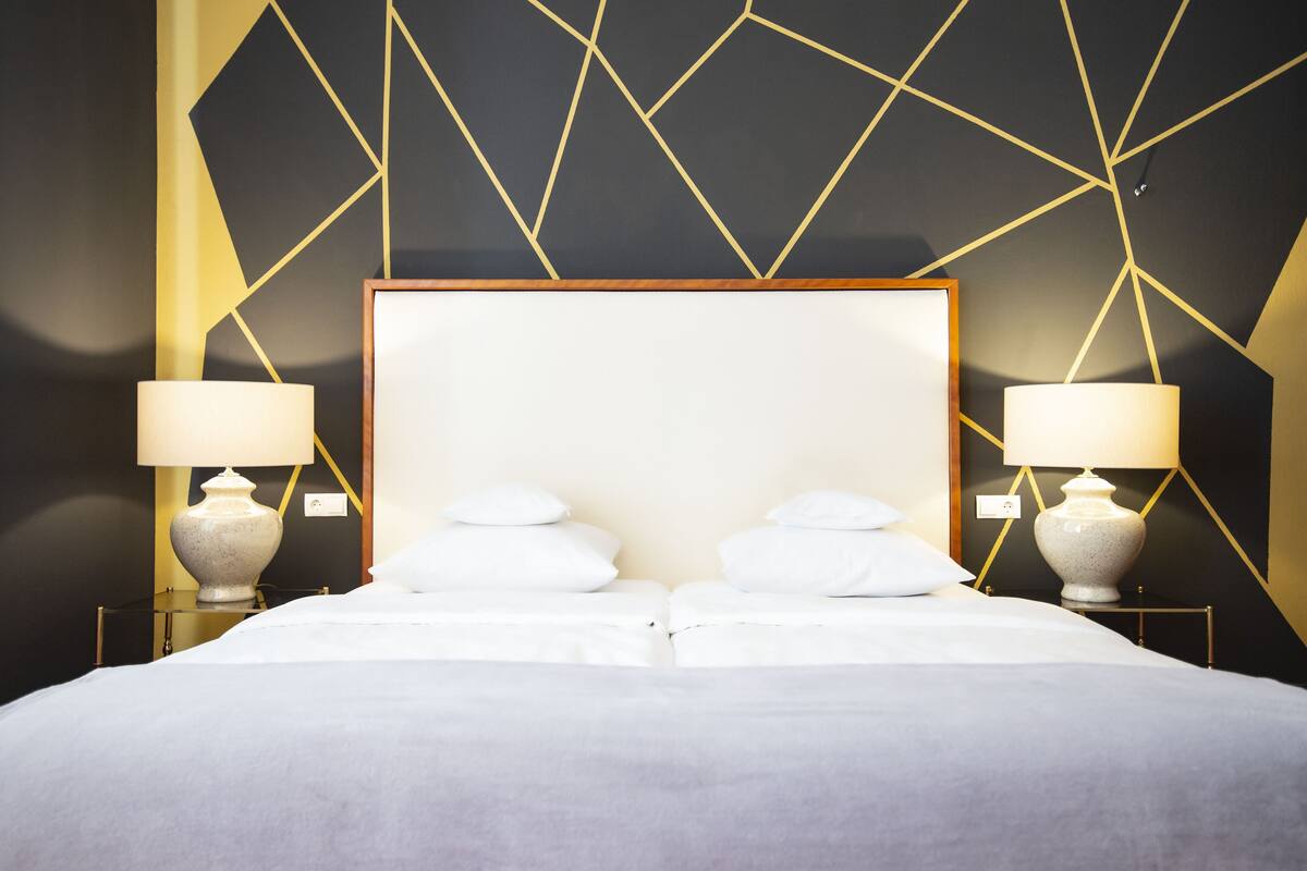 Uma cama de casal com cabeceira de madeira, duas mesas de cabeceira com luminárias e um design geométrico na parede.