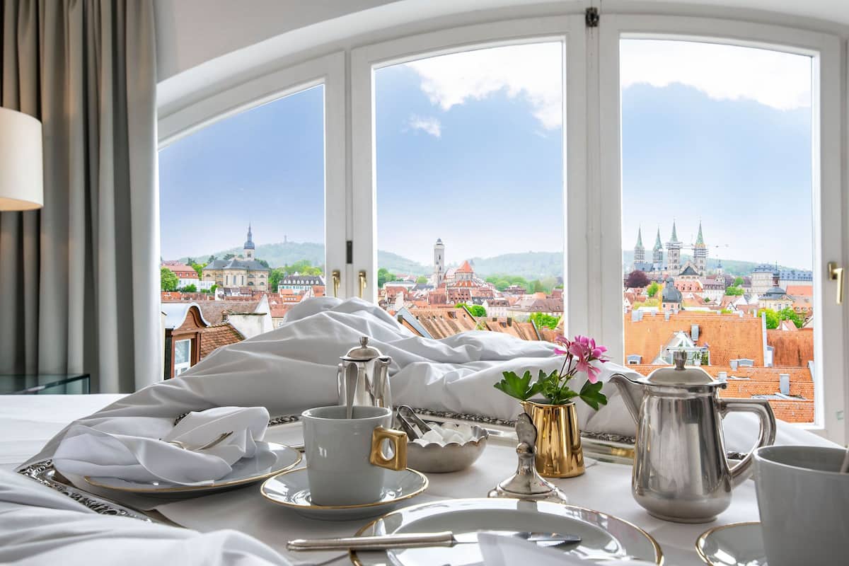 Mesa de café da manhã bem organizada com vista para um centro histórico através de grandes janelas.