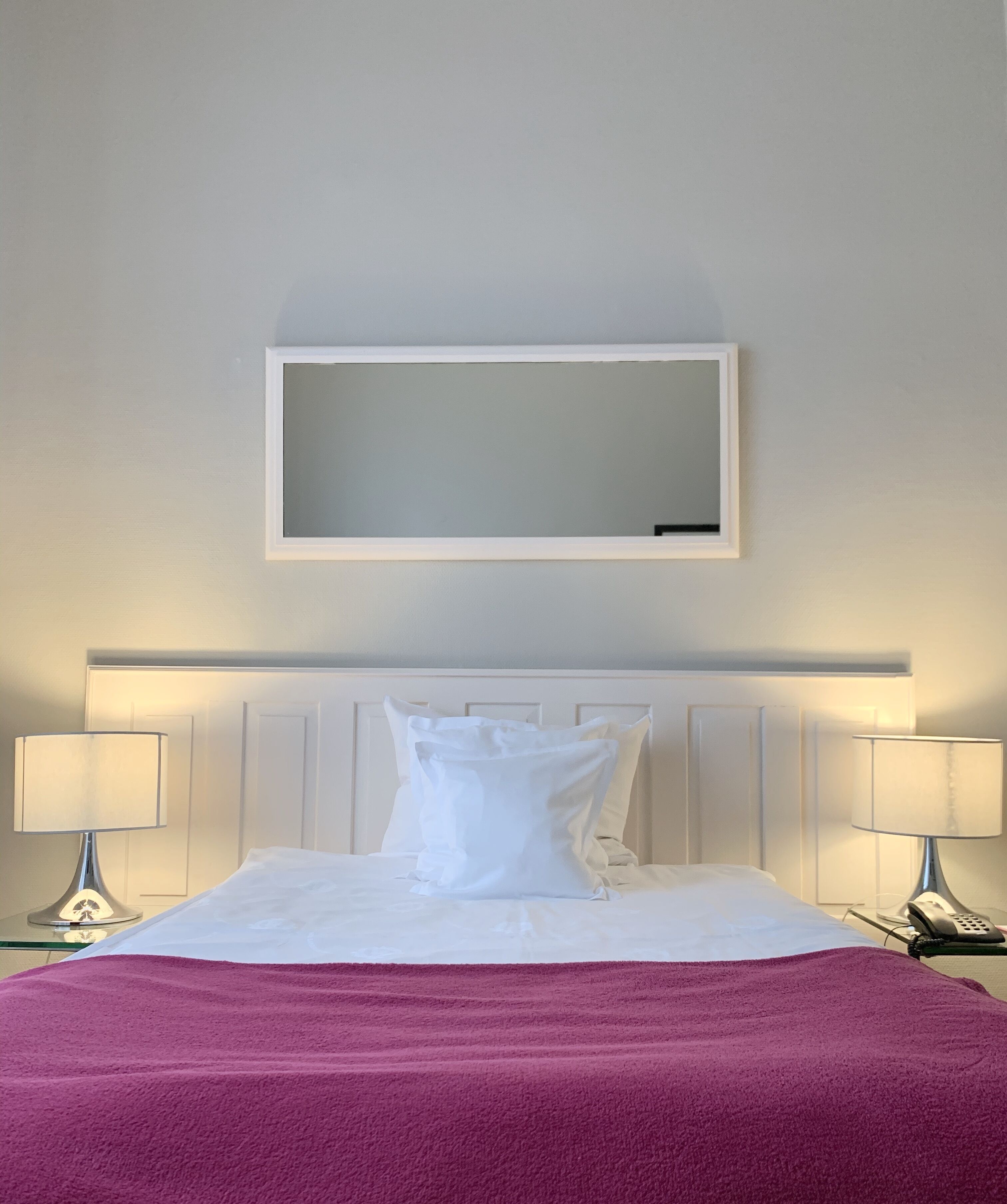 Une chambre d’hôtel avec un lit, deux lampes de chevet, un grand miroir au mur et un plaid rose.