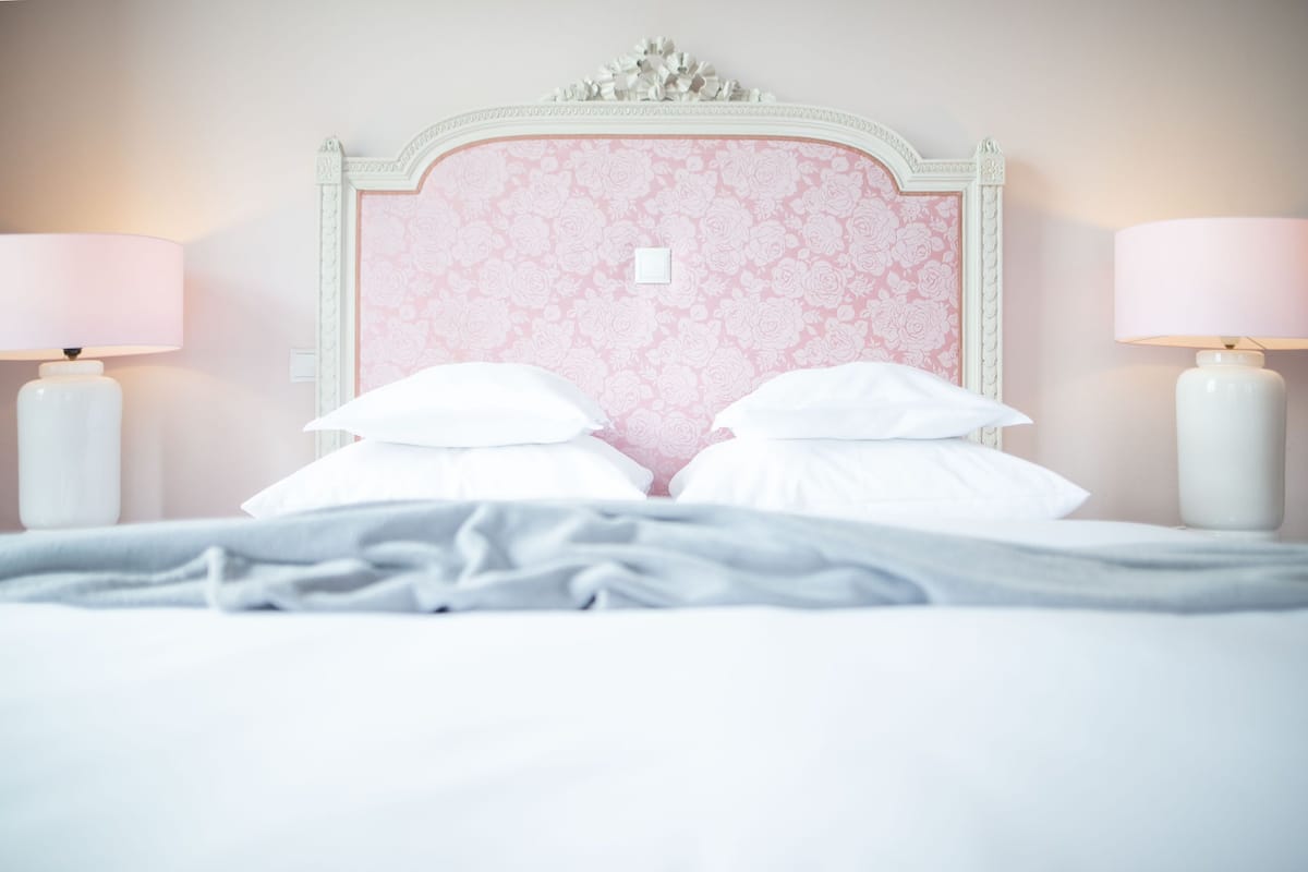Cama com lençóis brancos e cabeceira floral rosa. Duas luminárias de apoio de cada lado da cama.