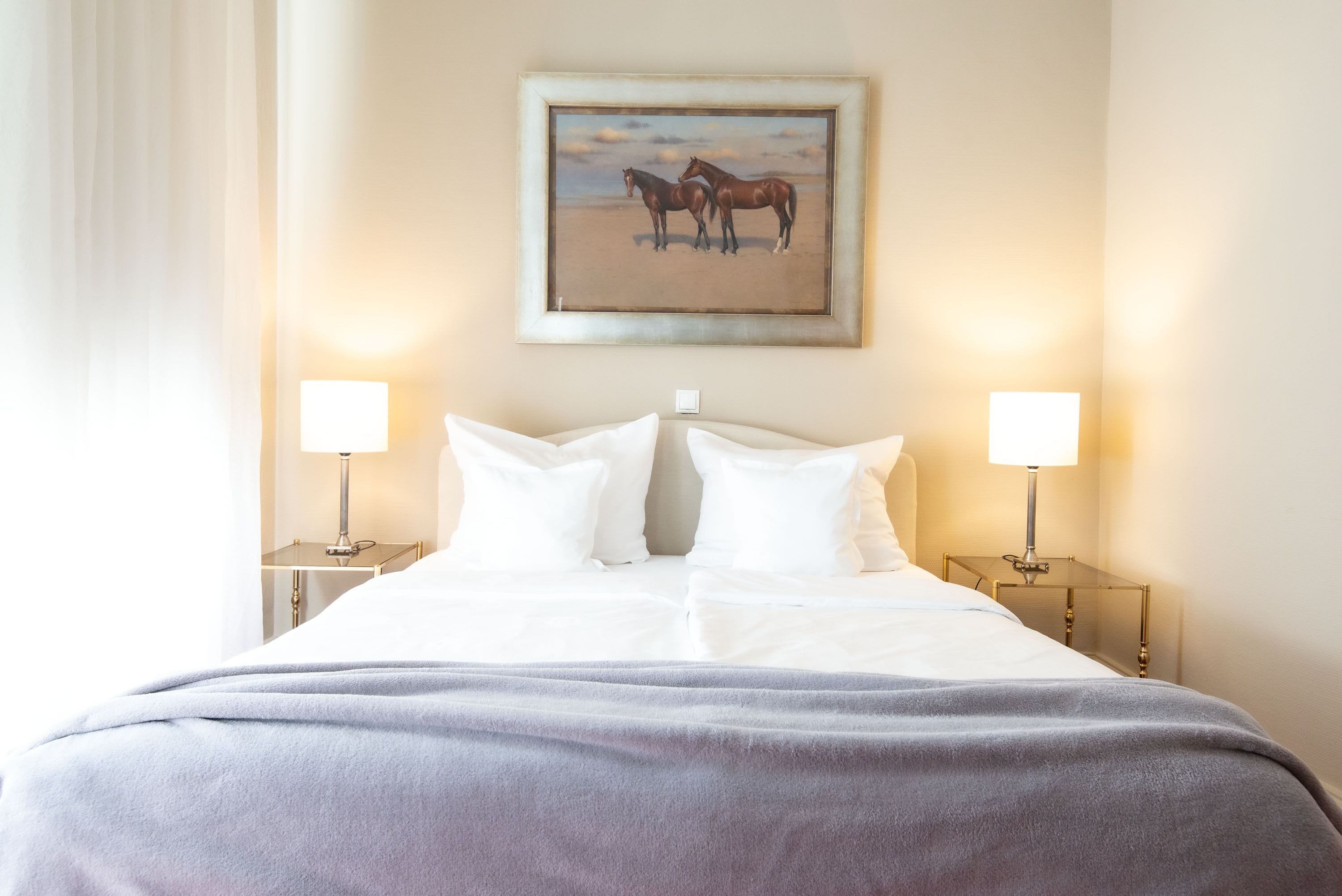 Habitación de hotel con una cama grande, dos lámparas de noche, un cuadro de caballos en la pared y una ventana con cortinas.