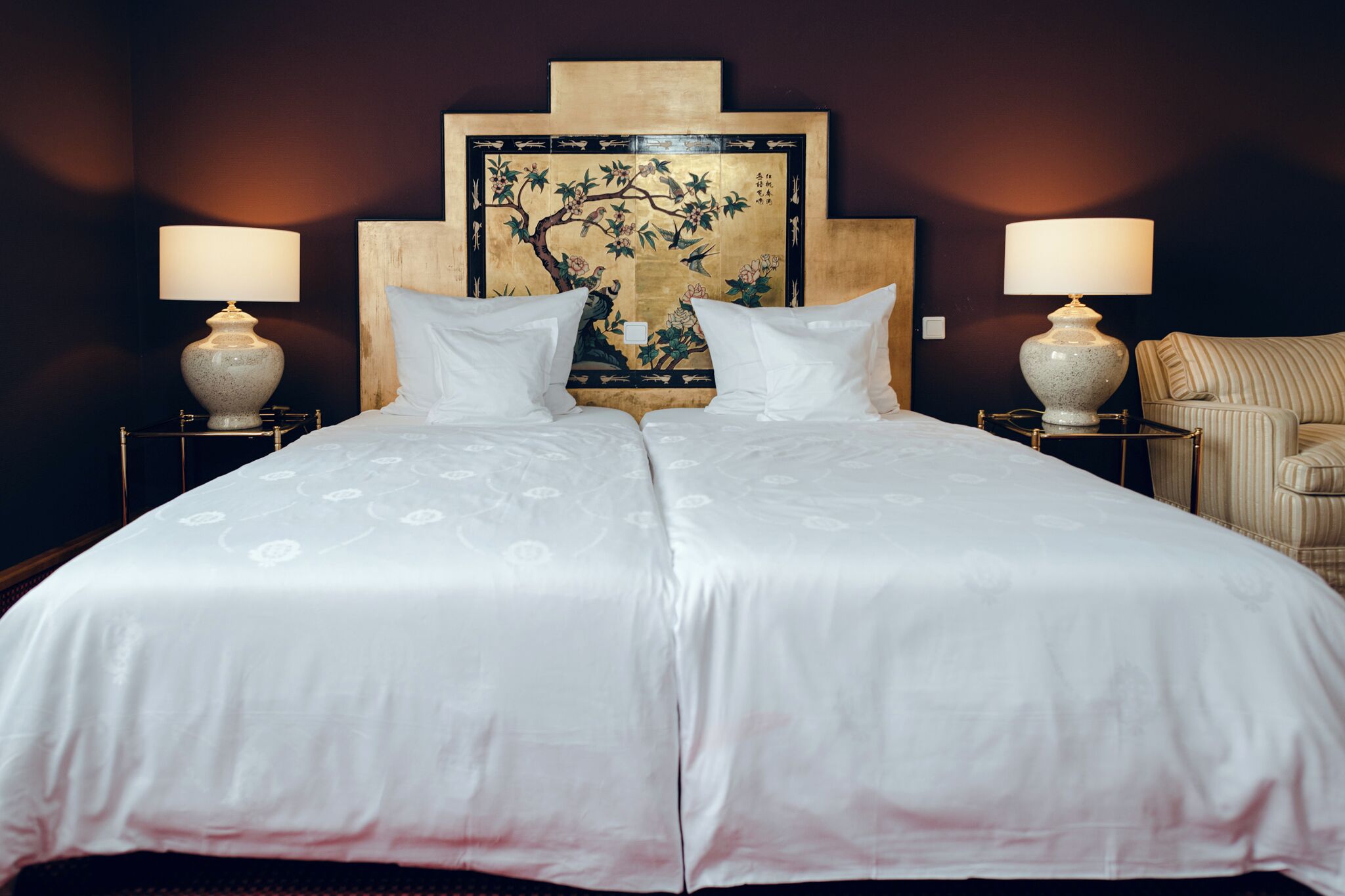 A double bed with white linens, two bedside tables with lamps, a large framed artwork on the headboard, and a striped armchair.