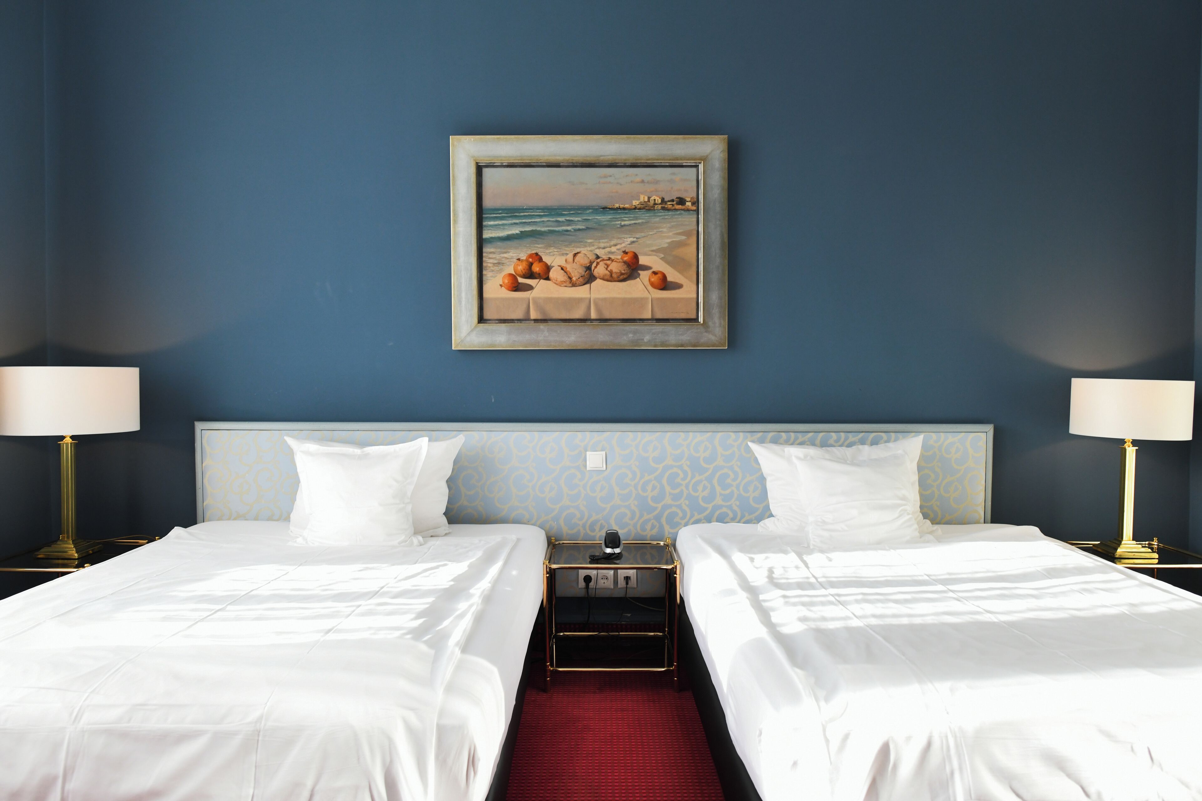 Une chambre d’hôtel avec deux lits, une moquette rouge, un tableau représentant une scène de plage et deux lampes de chevet.