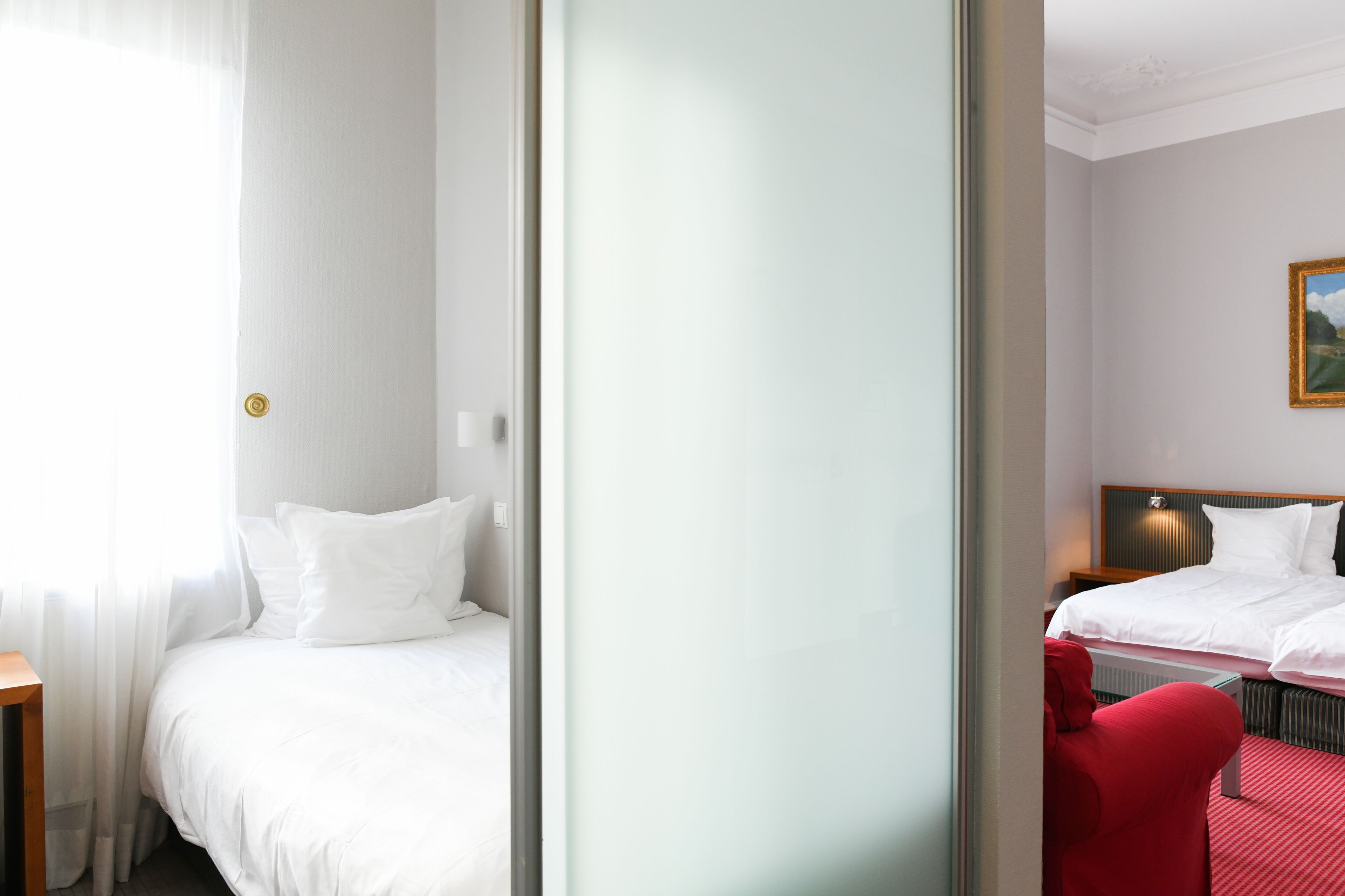 A hotel room with a bed, a red chair, and a painting on the wall.