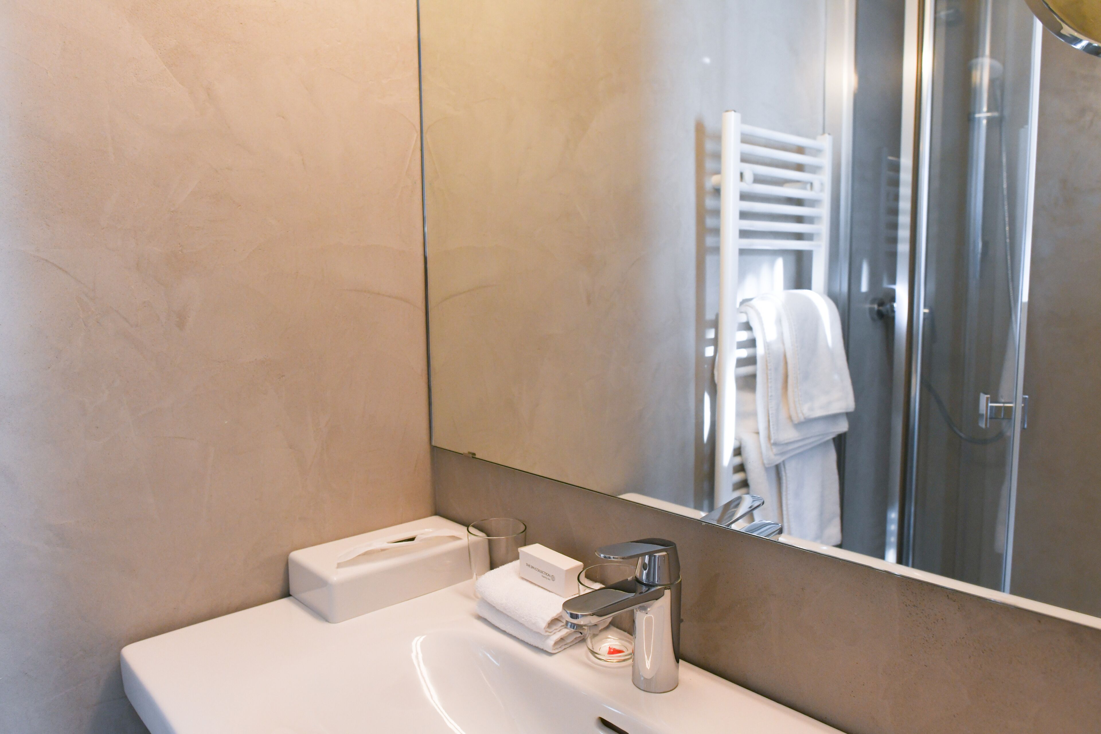 Ein modernes Badezimmer mit einem weißen Waschbecken, einem Glasbehälter, einem Handtuch und einem Spiegel, der einen Duschbereich reflektiert.