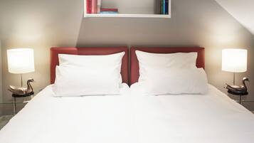 A neatly made bed with white linens and two pillows, flanked by two bedside tables with lamps and swan-shaped light fixtures. A bookshelf with books is mounted above the bed.