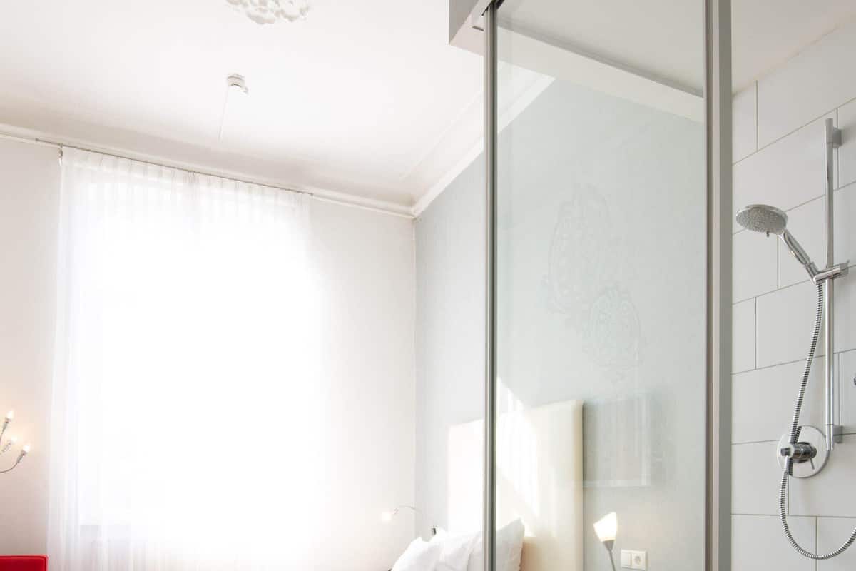 Quarto de hotel com cama, box de chuveiro de vidro, uma cadeira vermelha e piso de madeira.