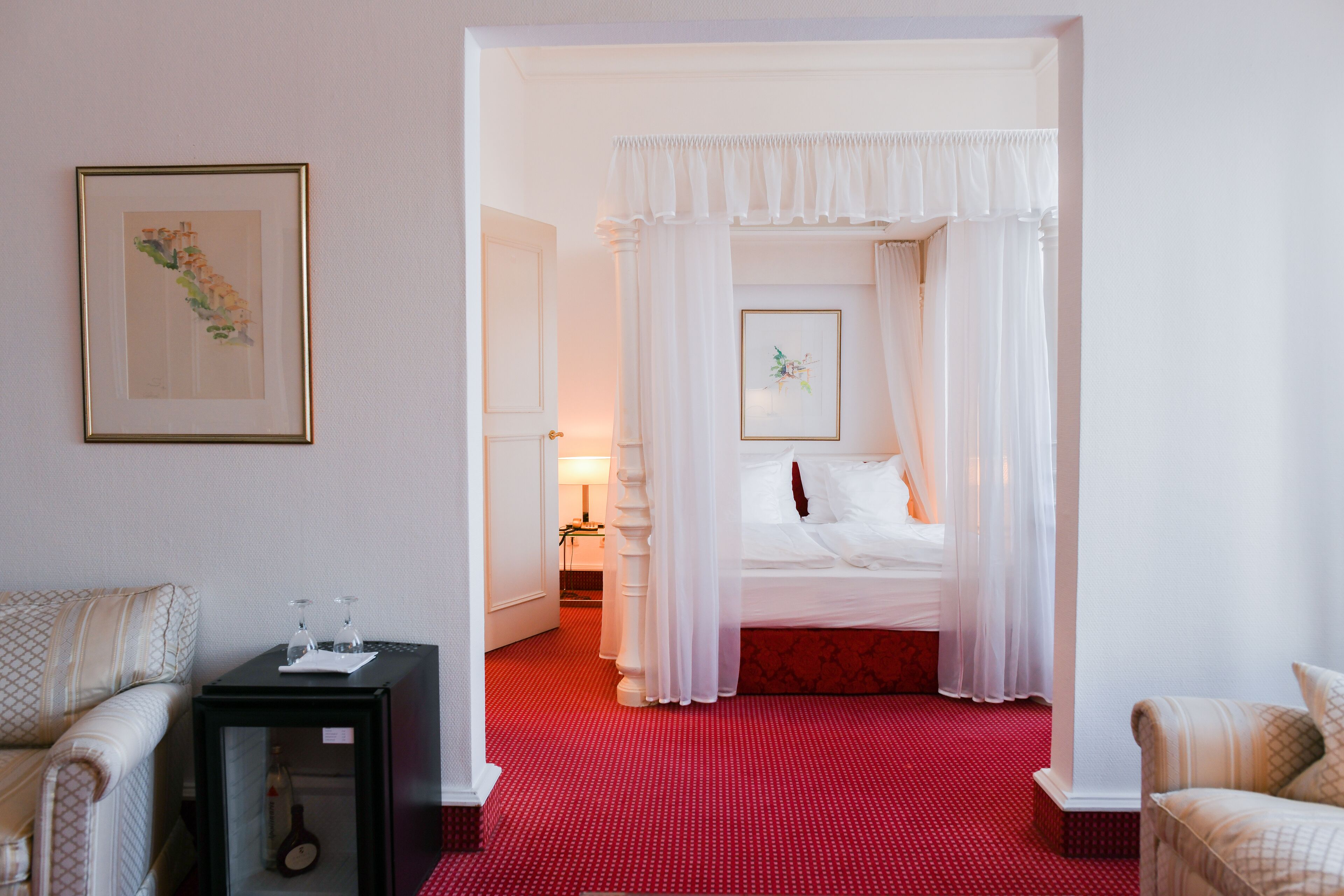 Habitación de hotel con alfombra roja, cama con ropa de cama blanca, mesita de noche con lámpara y un cuadro en la pared.