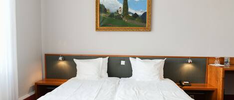 A hotel room with a large bed, two bedside tables, a nightstand, a framed painting on the wall, and a window with curtains.
