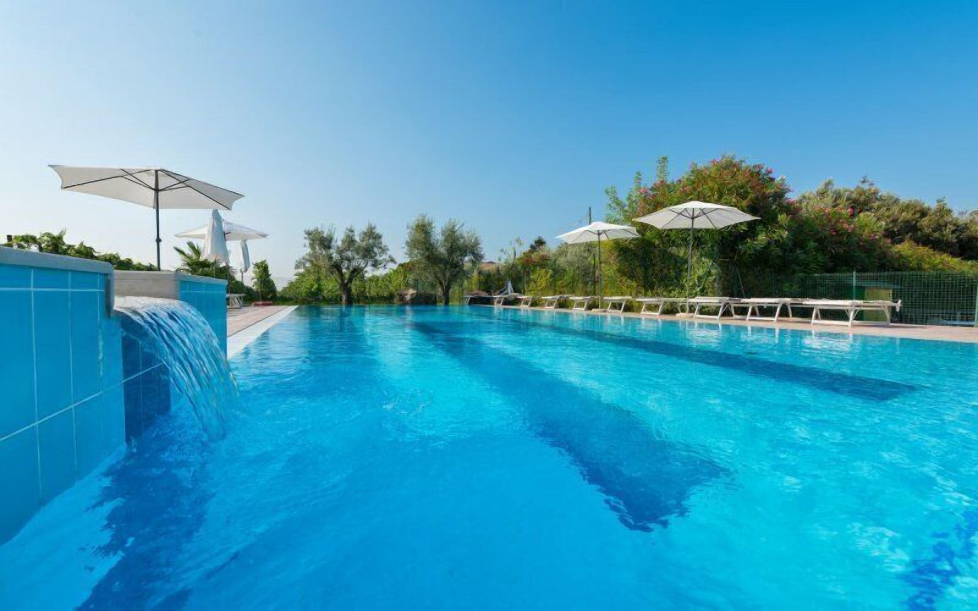 A clear blue swimming pool with a waterfall feature on the left, surrounded by lounge chairs and umbrellas.