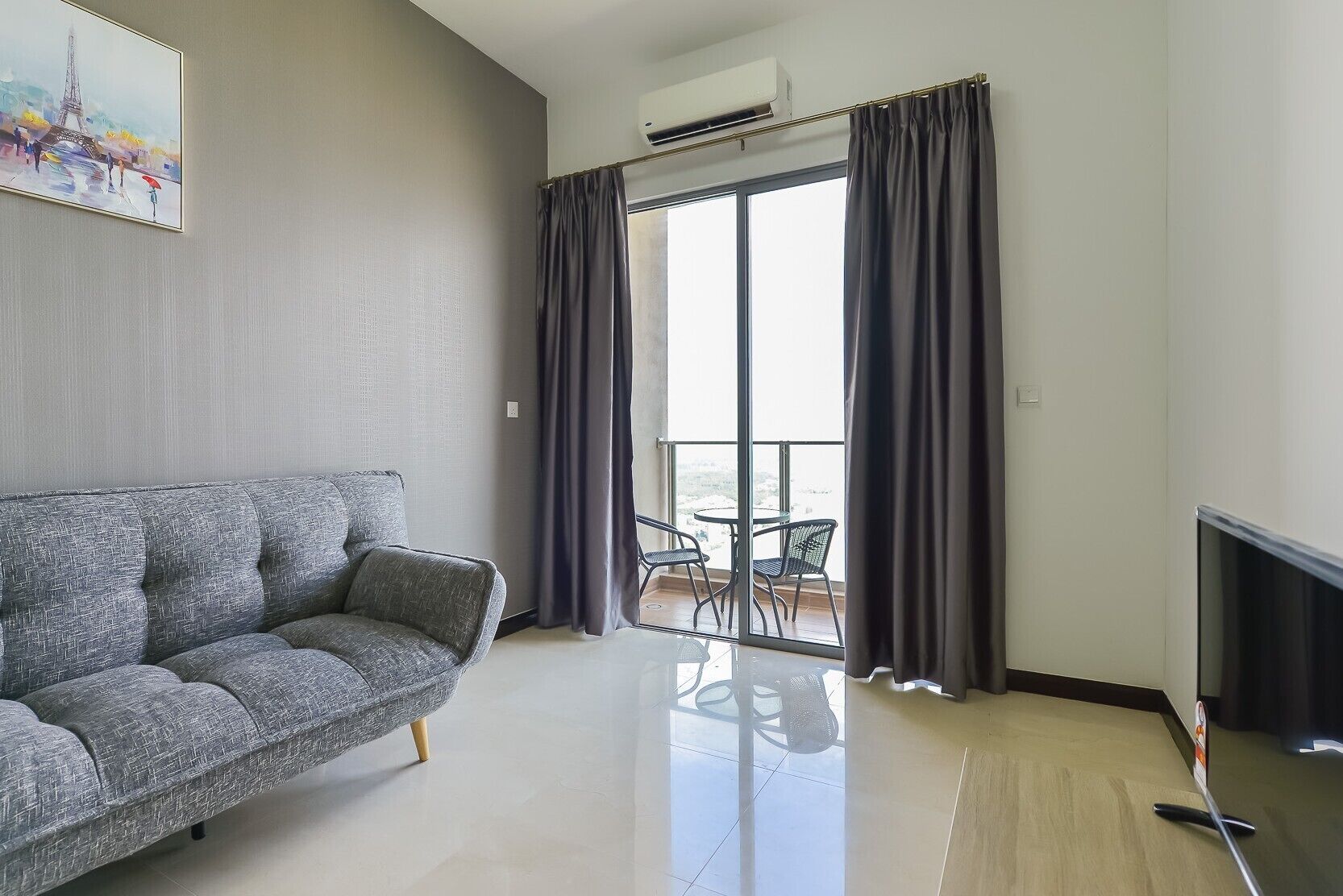 A modern living room with a grey sofa, a balcony with a table and chairs, and a painting of the Eiffel Tower.