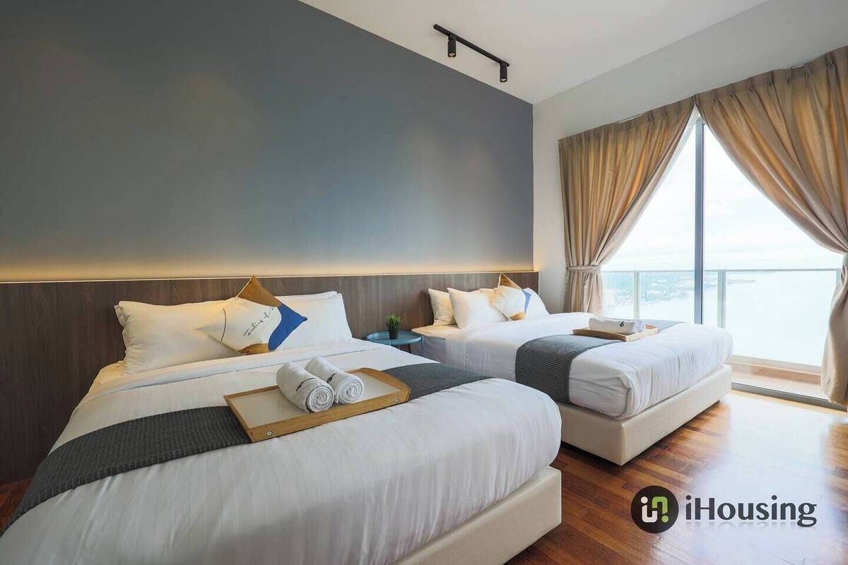 A hotel room with two beds, a large window with curtains, and a view of the sea.
