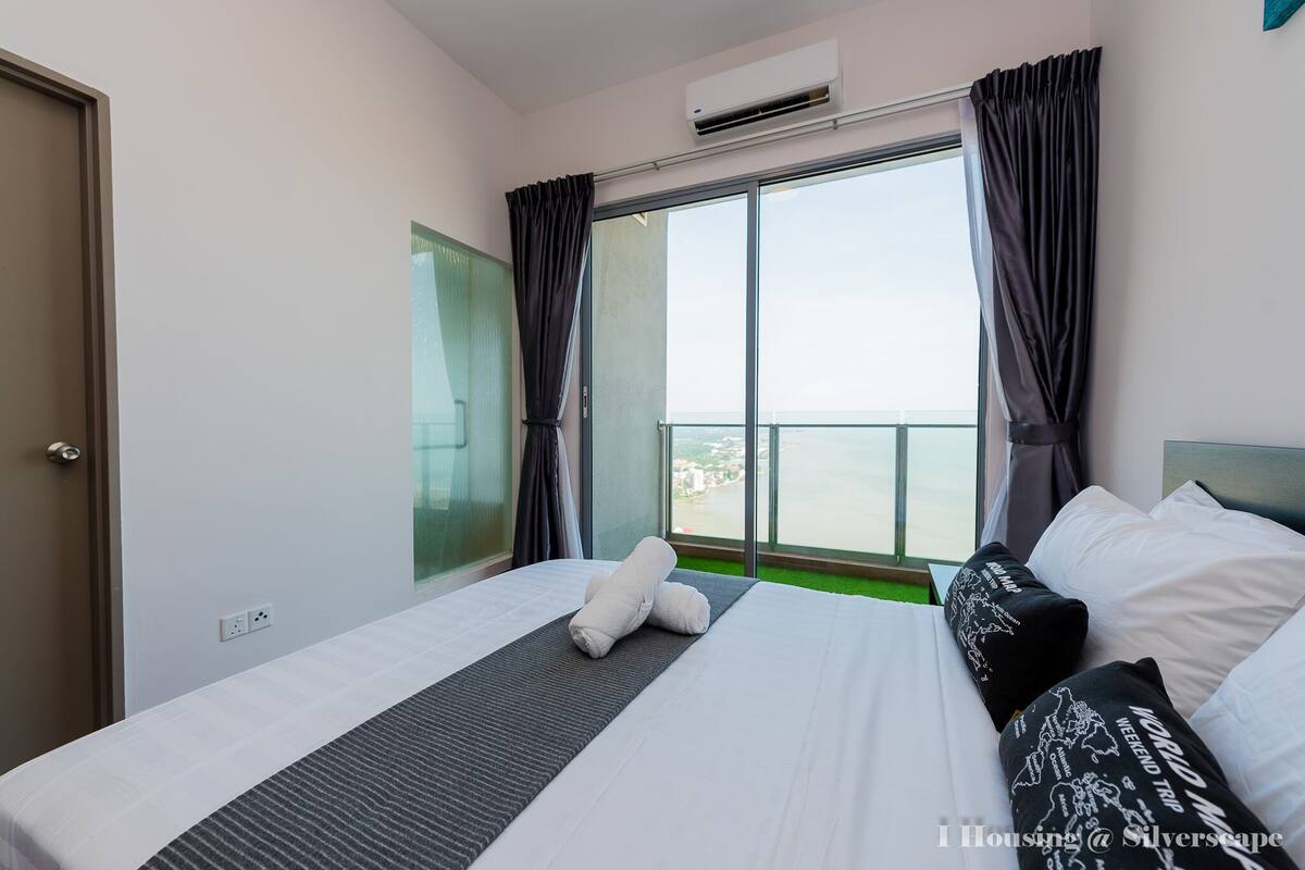 A hotel room with a large bed, a view of the sea through a sliding glass door, and a window with curtains.