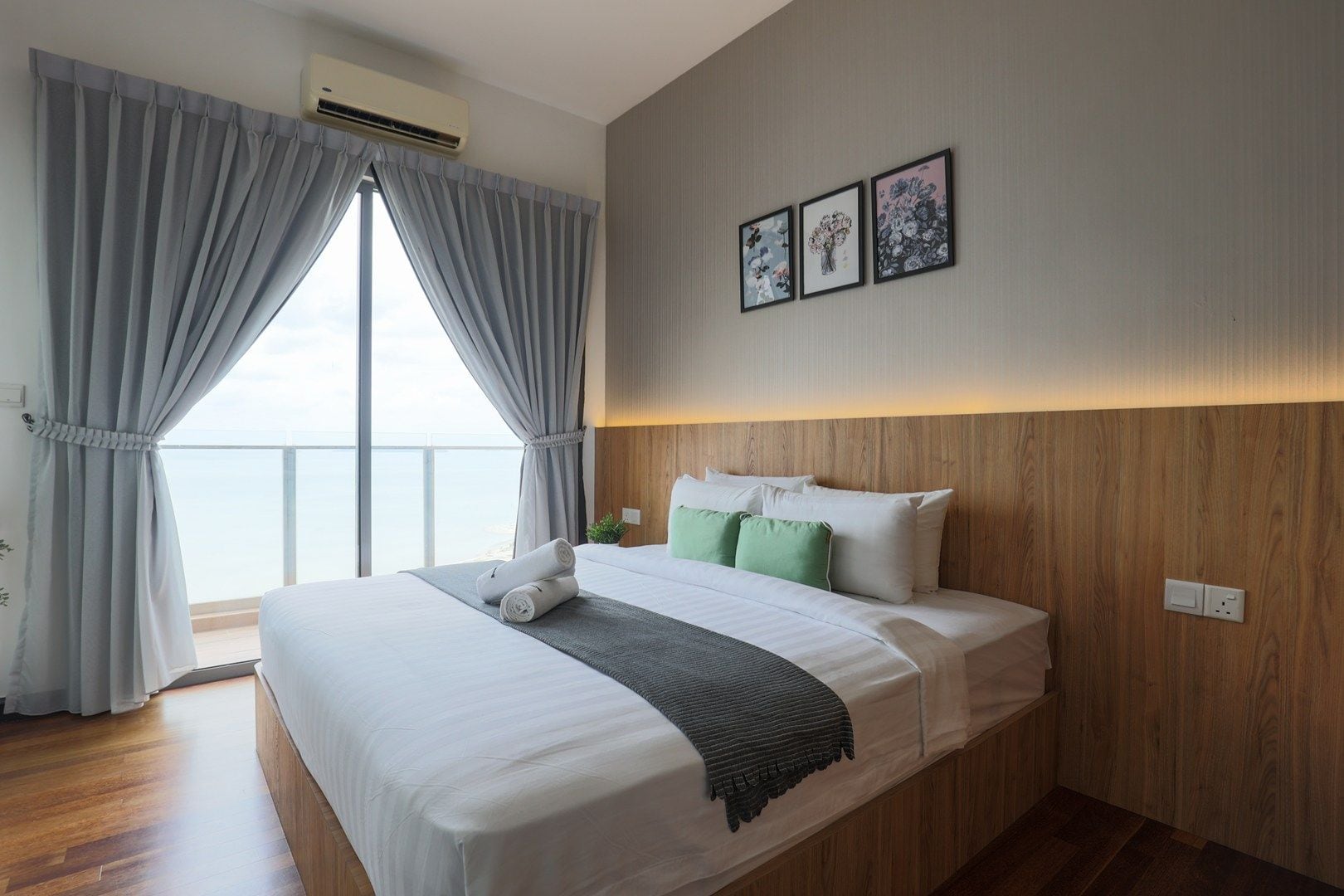 A hotel room with a large bed, grey curtains, a wooden headboard, and a view of the sea.