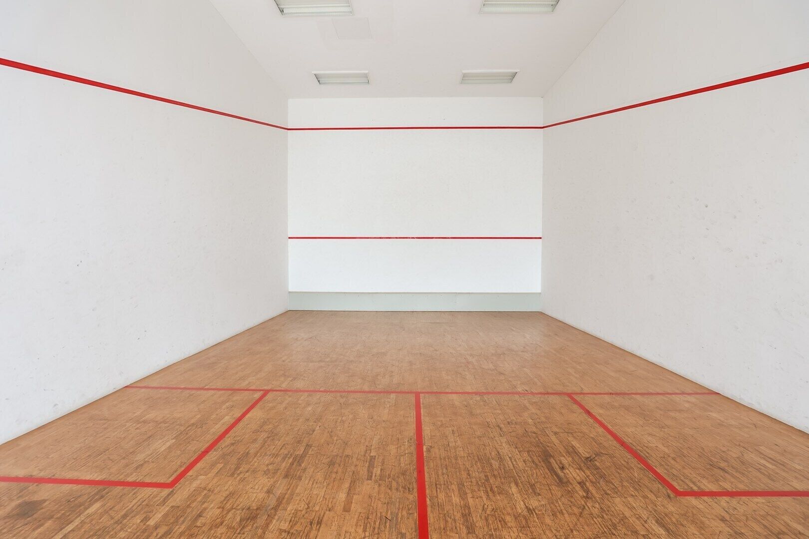 A squash court with white walls and a wooden floor marked with red lines.