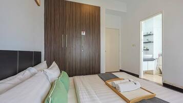 A hotel room with a bed, a wooden wardrobe, a bathroom visible through an open door, and a small wooden tray with towels.