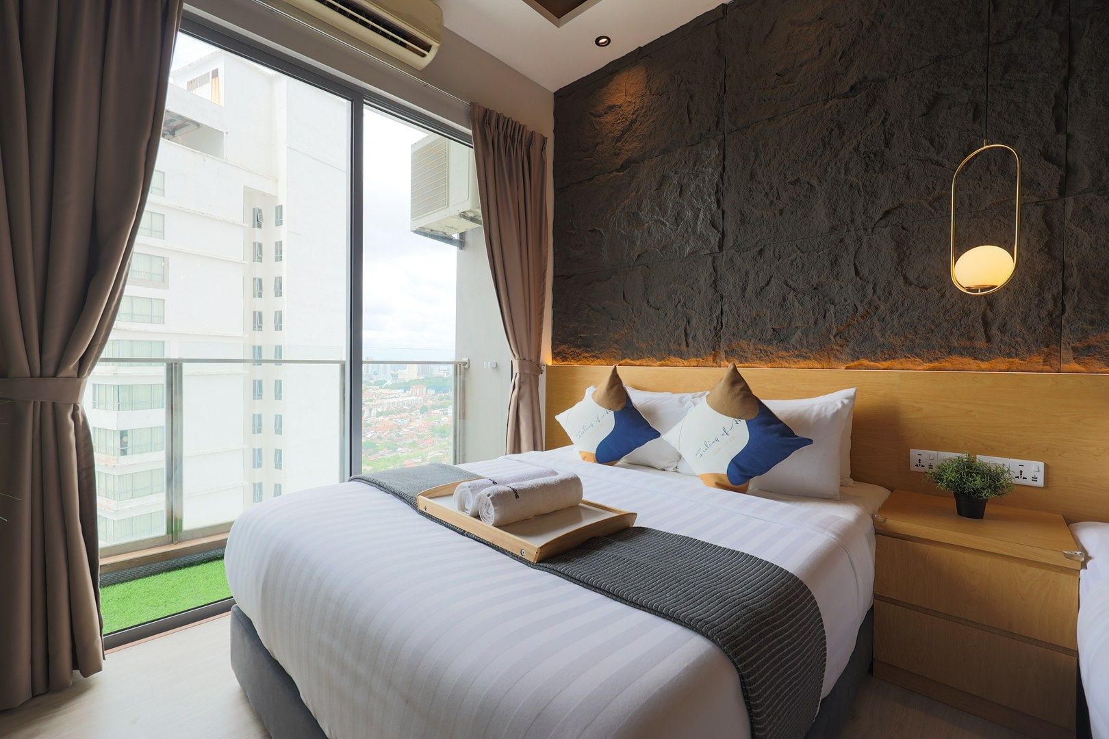 A hotel room with a large bed, a view of the city, a stone accent wall, and a bedside table with a plant.
