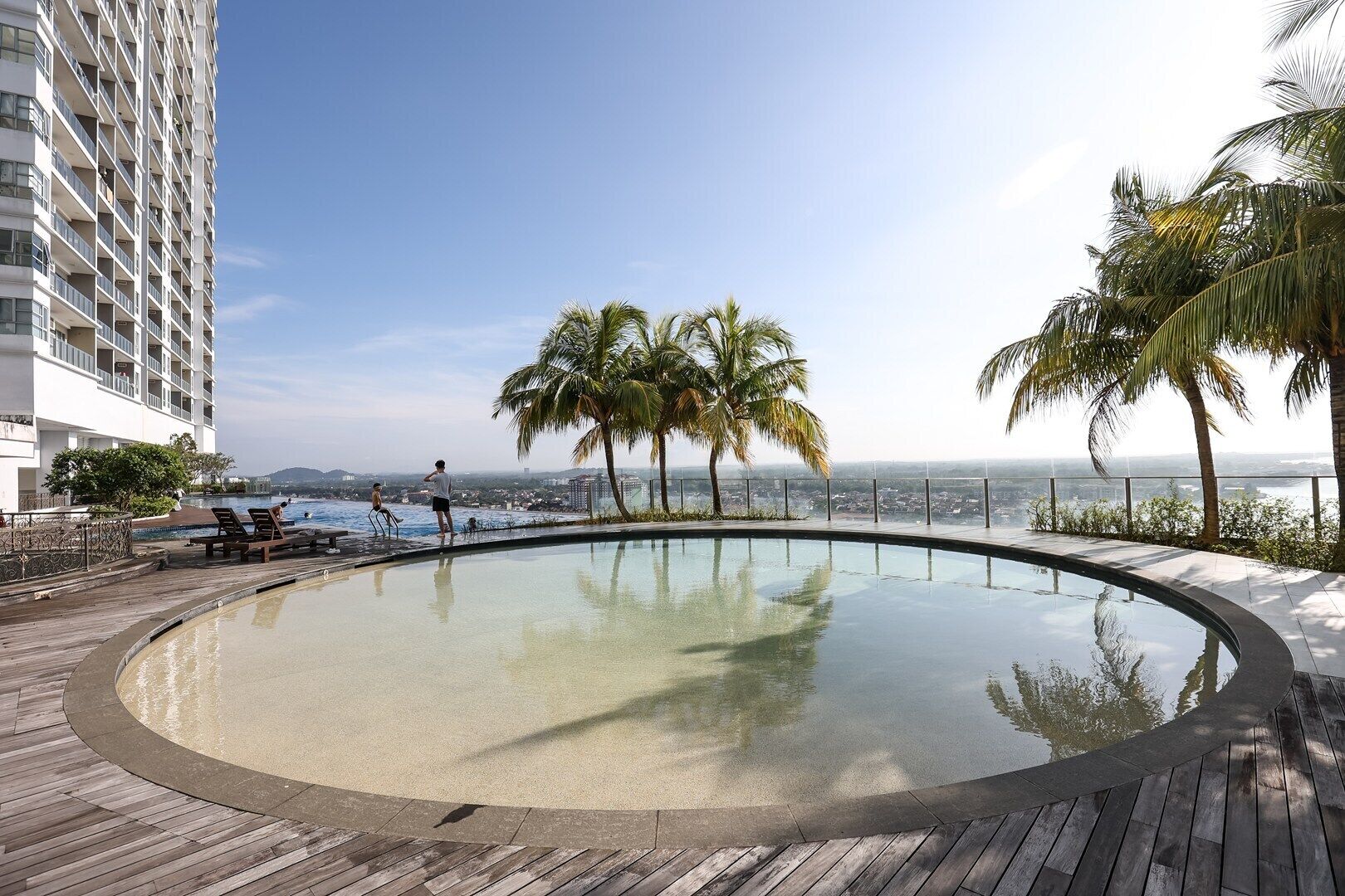 A rooftop infinity pool with a wooden deck, palm trees, and a view of the cityscape.