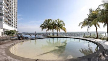 A rooftop infinity pool with a wooden deck, palm trees, and a view of the cityscape.