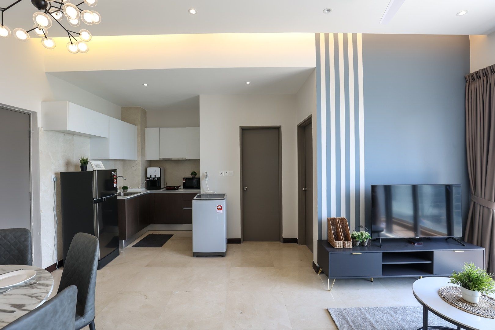 Uma sala moderna com televisão de tela plana, área de jantar com mesa de mármore e cozinha com armários escuros e geladeira.