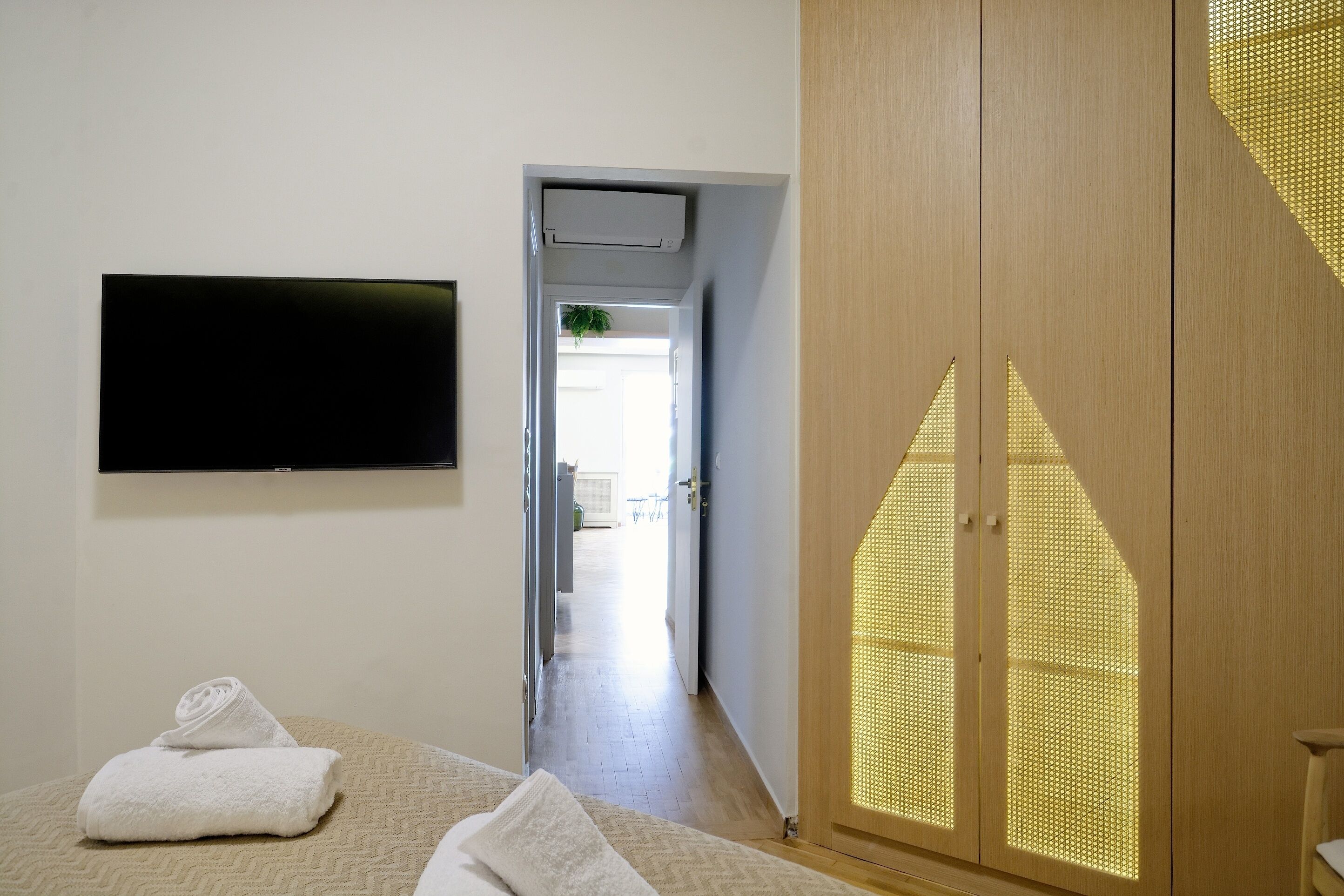 A hotel room with a bed, a TV mounted on the wall, a wooden wardrobe, and a hallway leading to a bathroom.