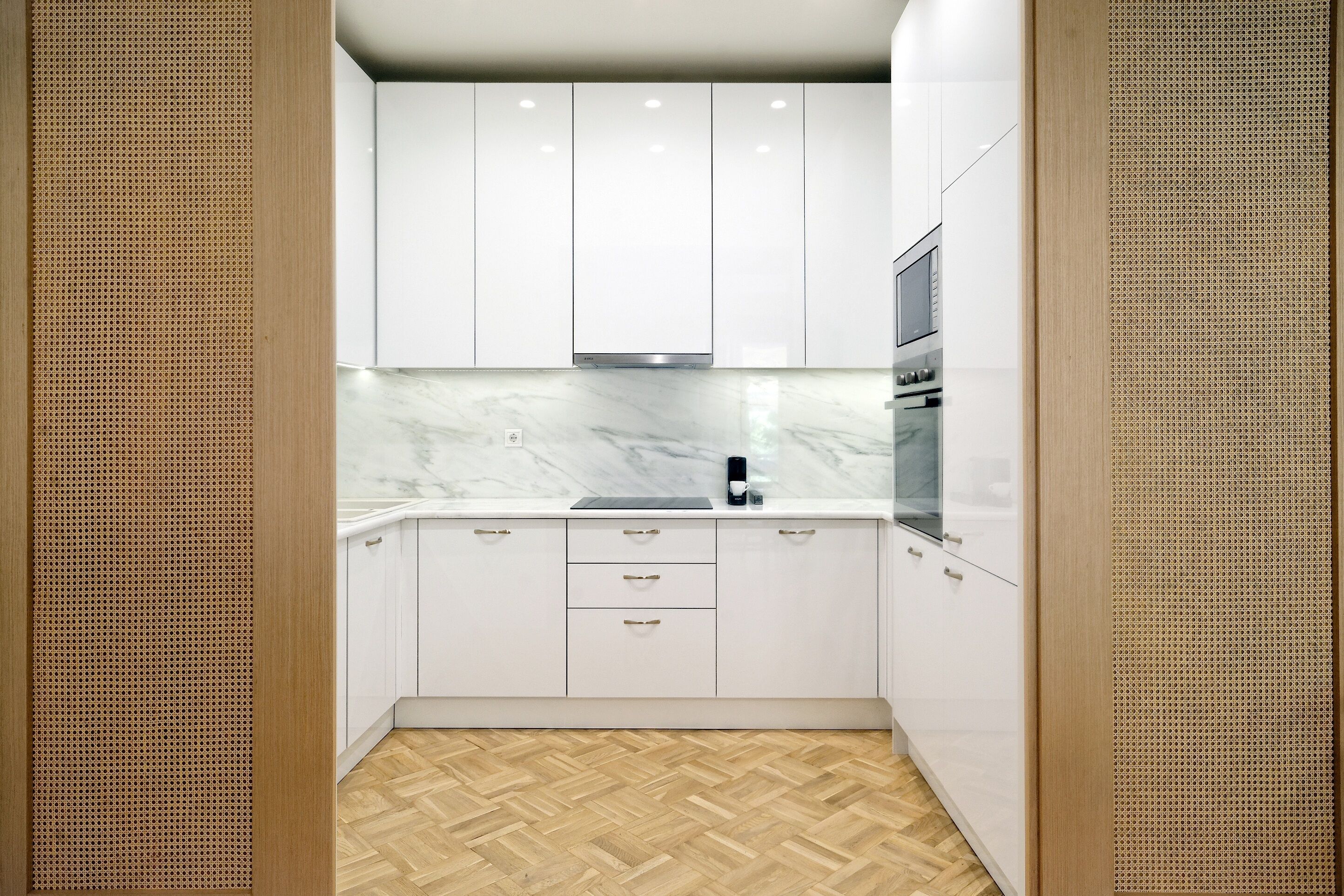 A modern kitchen with white cabinets, a marble countertop, and a herringbone wood floor.