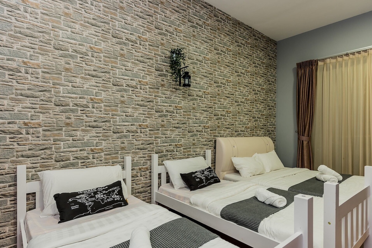 Una habitación con pared de piedra, dos camas con cabeceros blancos, una planta colgante y una ventana con cortinas.