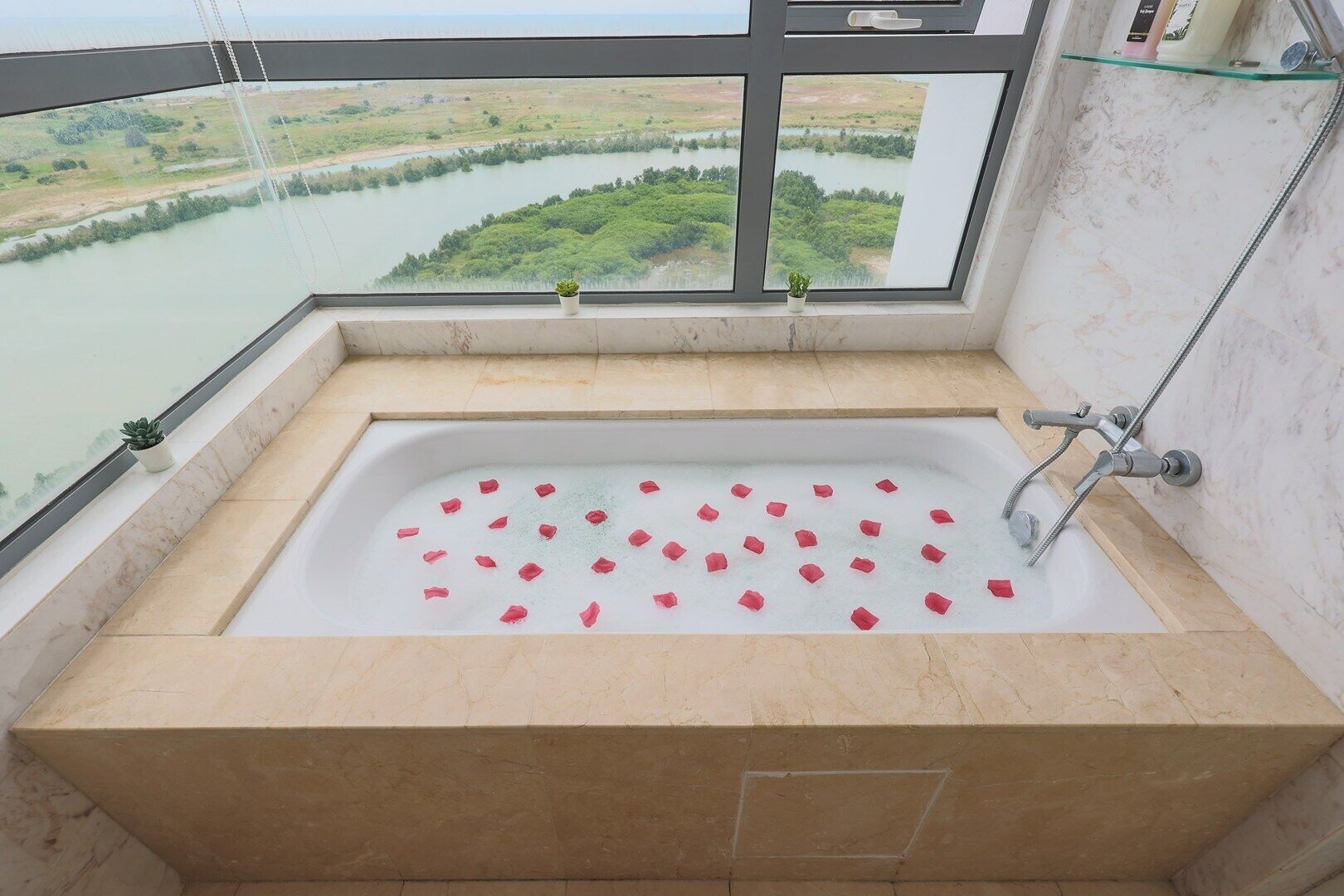 A bathtub with rose petals, a glass shelf with toiletries, and a view of a river and greenery.