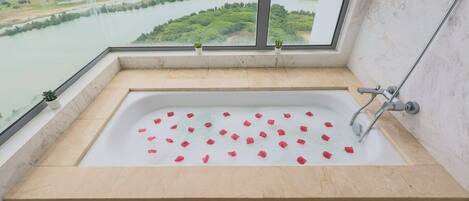 A bathtub with rose petals, a glass shelf with toiletries, and a view of a river and greenery.