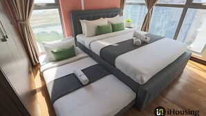 Una habitación de hotel moderna con una cama grande, una cama más pequeña y una cama sofá. La habitación tiene ventanas amplias con vistas a edificios y zonas verdes.