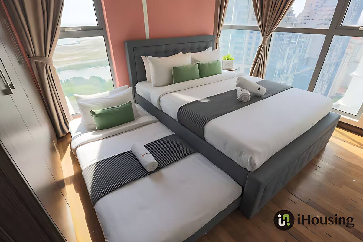 A modern hotel room with a large bed, a smaller bed, and a sofa bed. The room has large windows with a view of buildings and greenery.