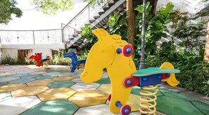A playground with colorful play structures, including a yellow horse and a blue dinosaur, set on a patterned tile floor with greenery in the background. - Silverscape Executive Melaka by I Housing (Malacca City)