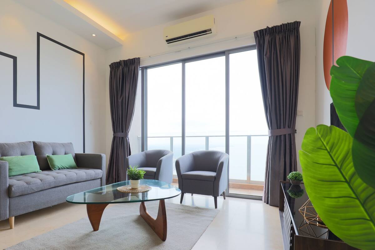 A modern living room with a grey sofa, two armchairs, a glass coffee table, and a large window with curtains.