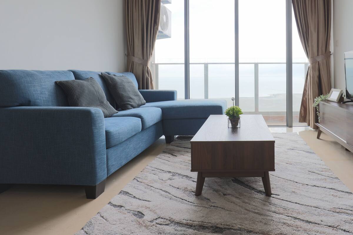 A modern living room with a blue sofa, a wooden coffee table, and a view of the sea through large windows.