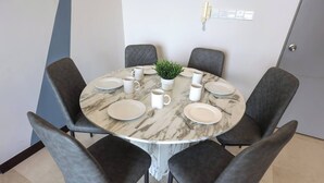 Una mesa de comedor redonda con seis sillas grises tapizadas, una planta en maceta, platos, tazas y cubiertos blancos.